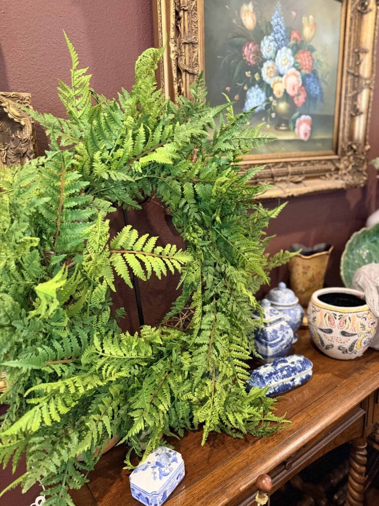 A green fern wreath sits on a wooden table decorated with blue and white ceramic jars and a patterned vase, beneath a framed floral painting on a burgundy wall.