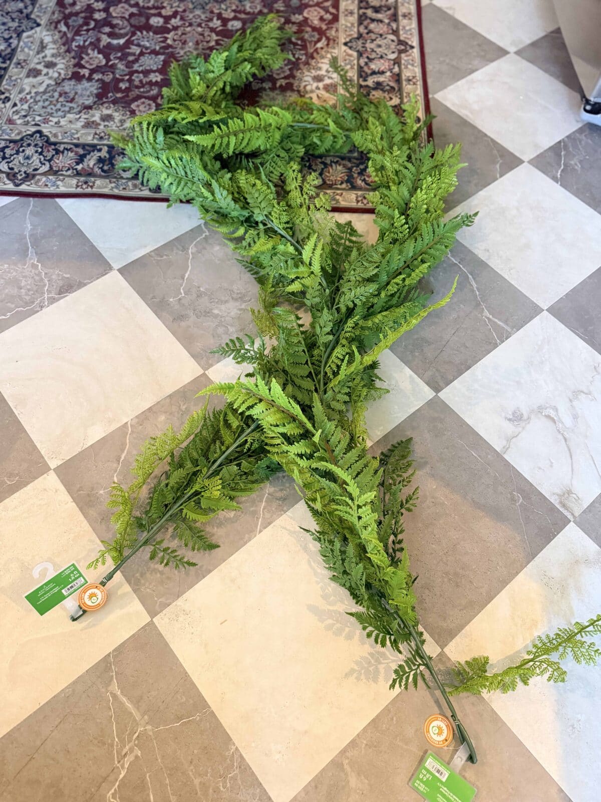 Two long artificial fern garlands with tags attached are laid out on a checkered tile floor near the corner of a patterned rug.