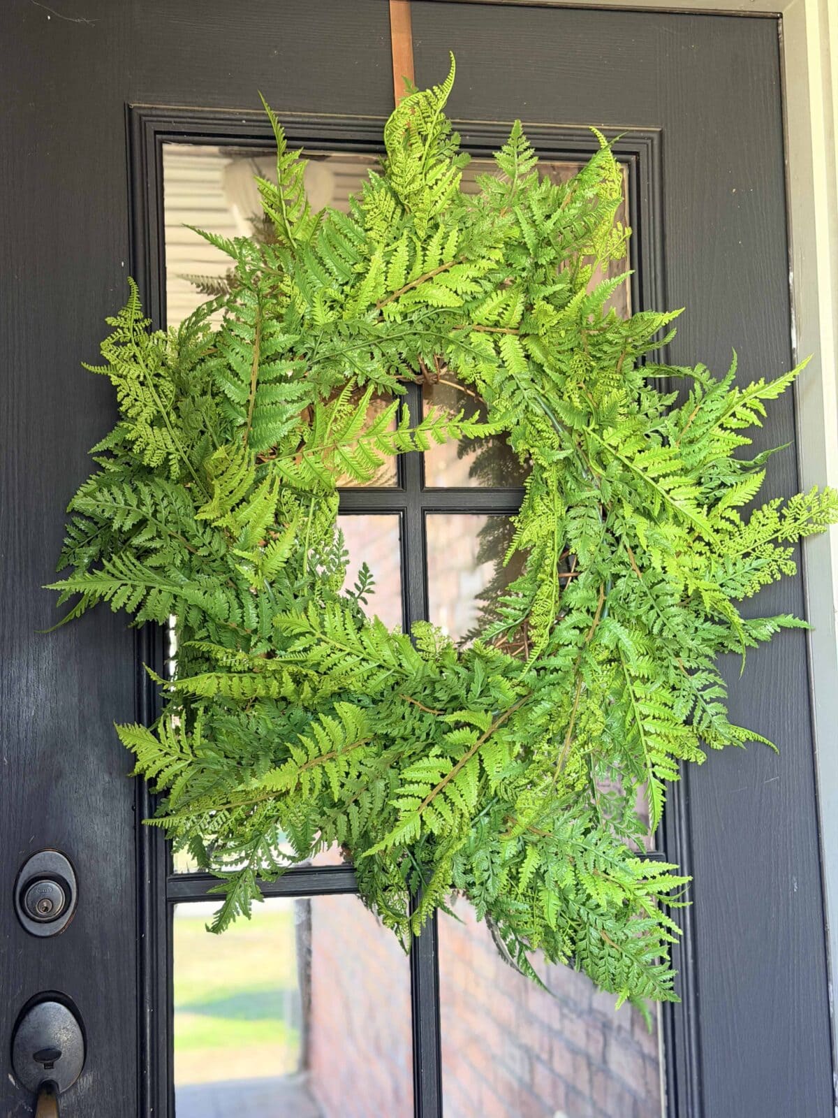 A vibrant green fern wreath hangs on a black front door with glass panels, secured by a brown ribbon. Sunlight filters through the window, and part of a porch and brick wall are visible outside.