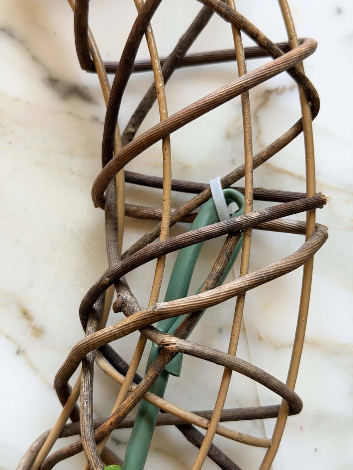 Close-up of two intertwined, twisted wicker strands in light and dark brown, with a green plastic stick and a white zip tie intertwined among them, all resting on a cream-colored marble surface.