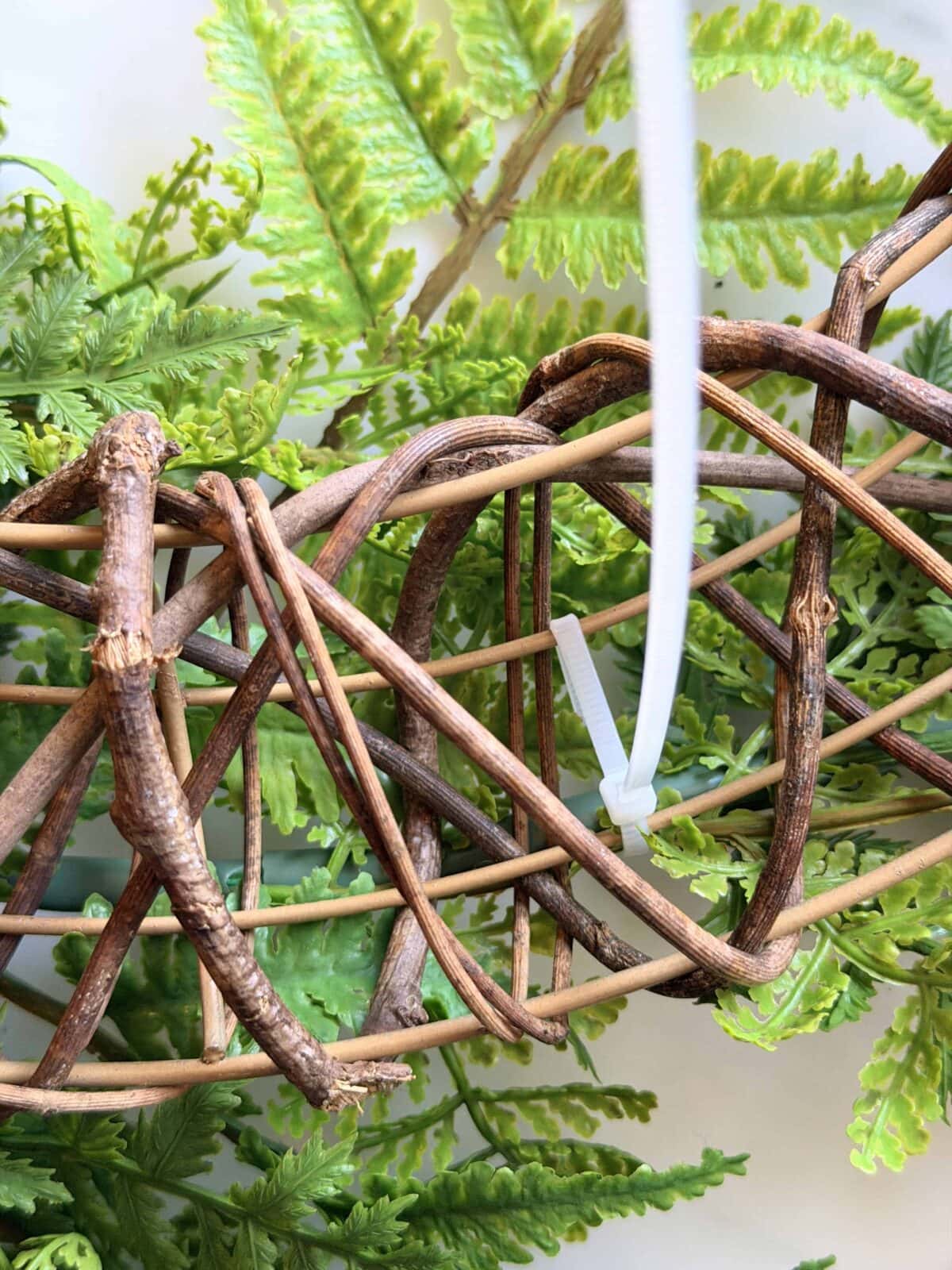 A close-up of intertwined brown wicker vines with green artificial ferns in the background. A white plastic zip tie is attached to the wicker structure.
