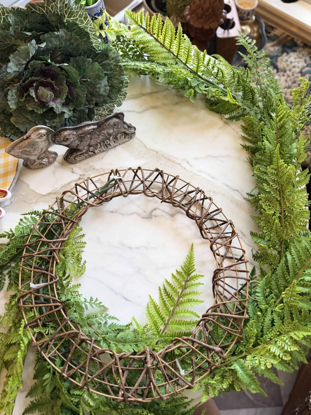 A grapevine wreath forms the base on a marble surface, surrounded by bright green fern fronds. Decorative items, including a cabbage and ceramic rabbit, are visible in the background.