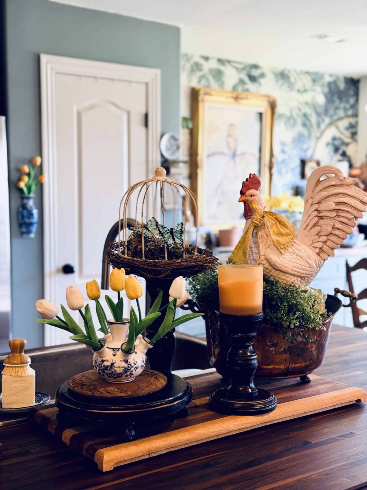 A kitchen table styled for a spring home tour features white tulips in a teapot, a yellow candle on a stand, a birdcage with twigs and moss, and a ceramic rooster with a yellow ribbon, all set against a cozy kitchen backdrop.