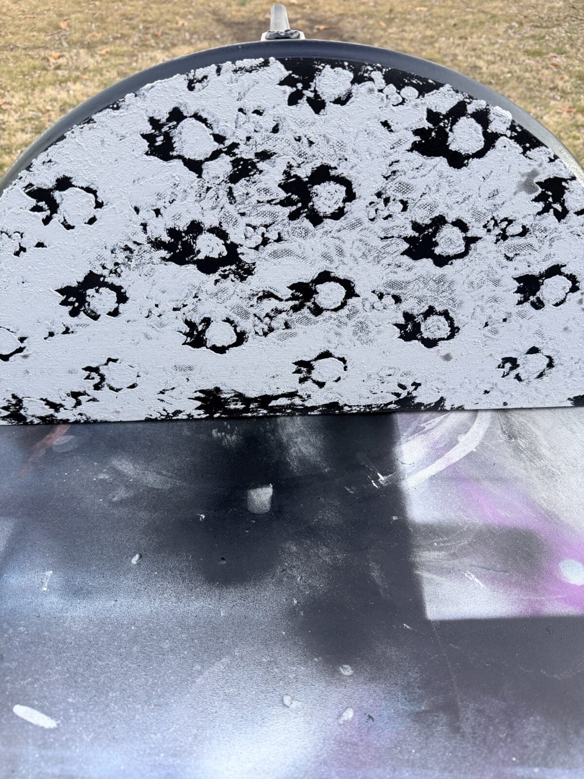 A close-up of a curved, half-circle metal surface painted with black and white floral or splatter patterns, set outdoors on grass with cloudy sky reflections visible on the lower section.