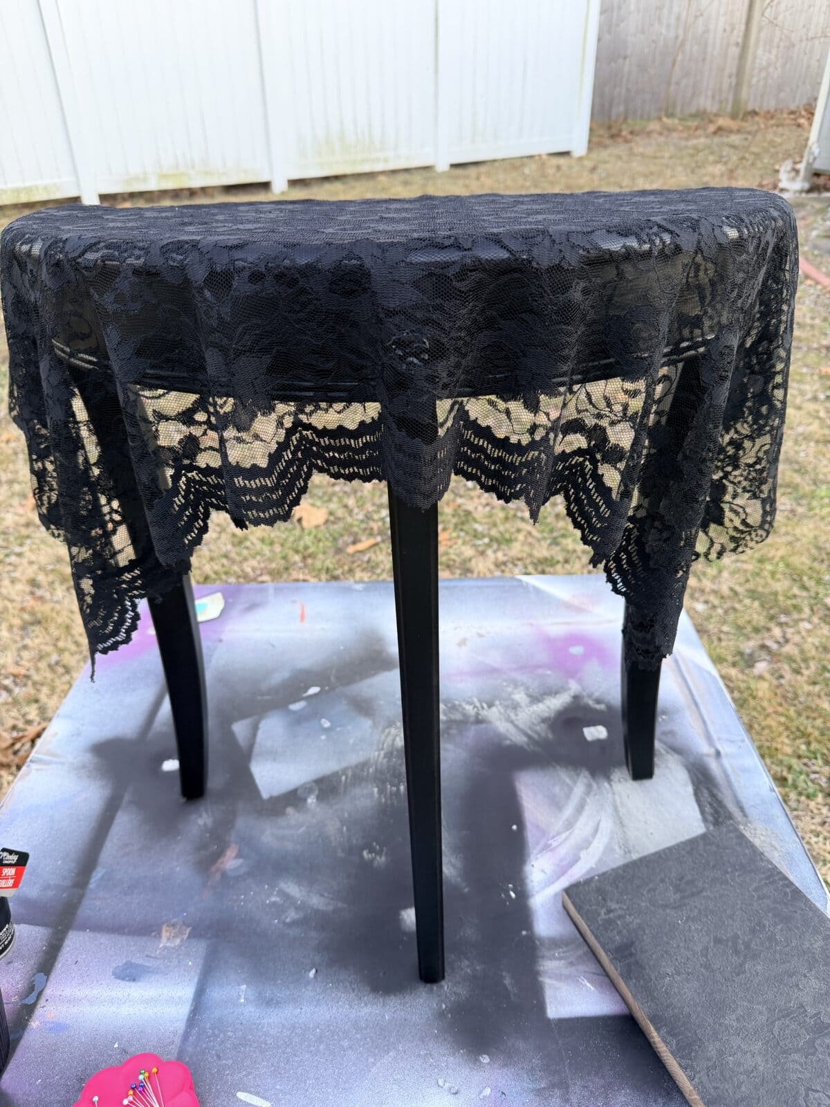 A small round table stands upside down outdoors on a spray-painted surface, covered with a black lace cloth. The background shows grass, a wooden fence, and white fencing panels.