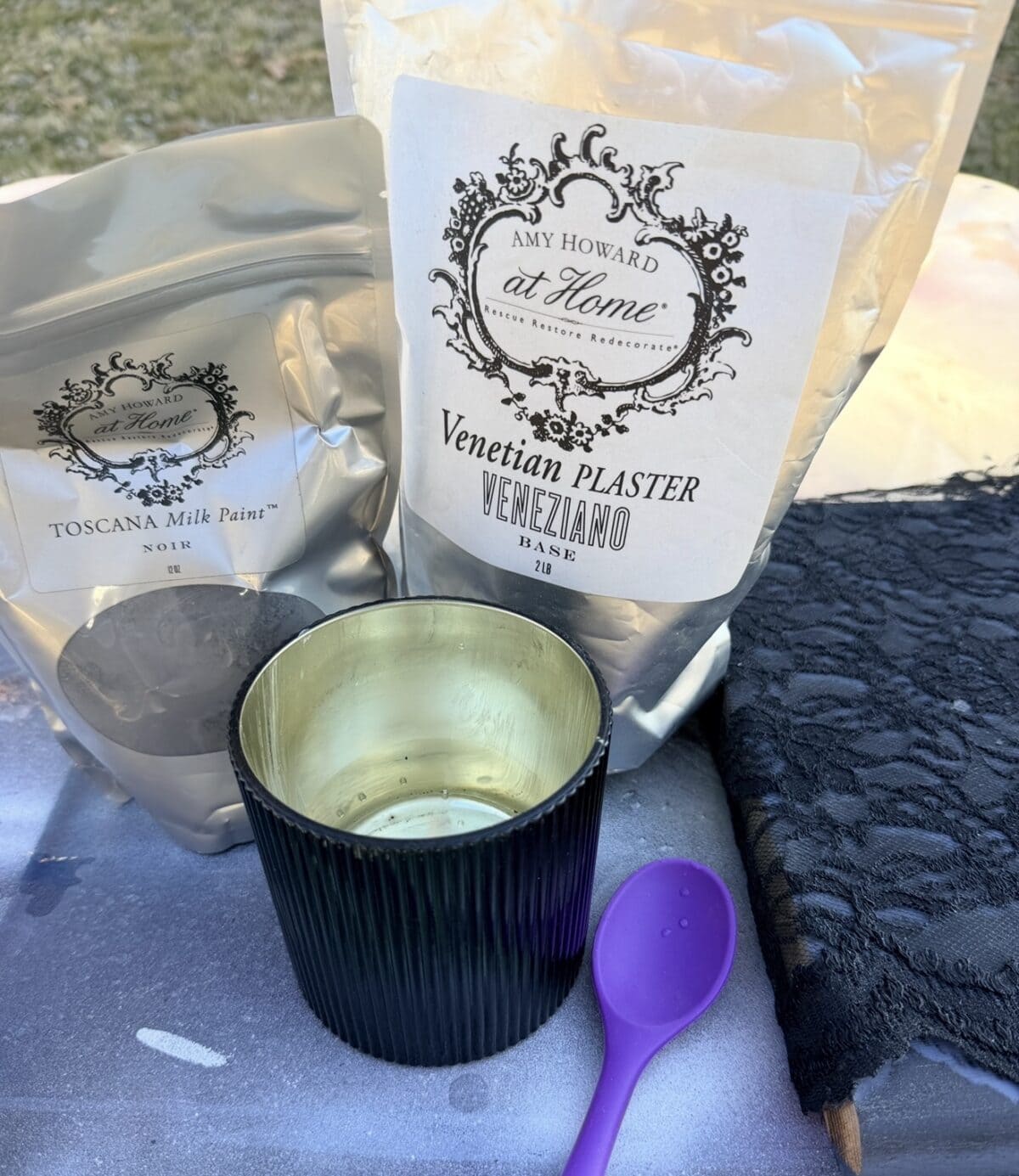 A black and gold candle holder, a purple spoon, black lace fabric, and two bags labeled "Toscana Milk Paint" and "Venetian Plaster" from Amy Howard at Home are displayed on a table outdoors.