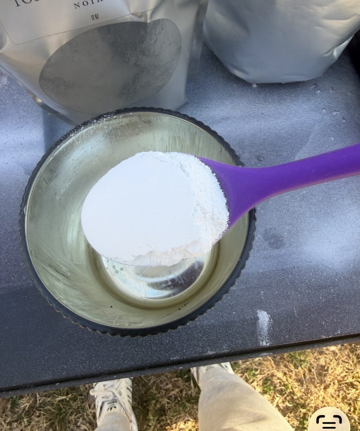 A purple spoon holds a heaping scoop of white powder over a glass bowl. The scene is set outdoors on a black table, with someone's white shoes and grass visible below. Nearby, there are gray bags in the background.