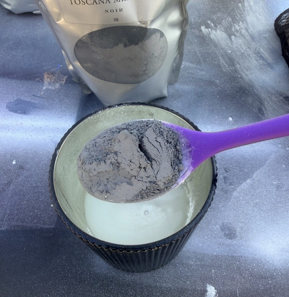 A purple spoon holds a heap of gray powder over a cup filled with white liquid. In the background, a silver bag labeled "Toscana" contains more of the gray powder, some of which is scattered on the surface.