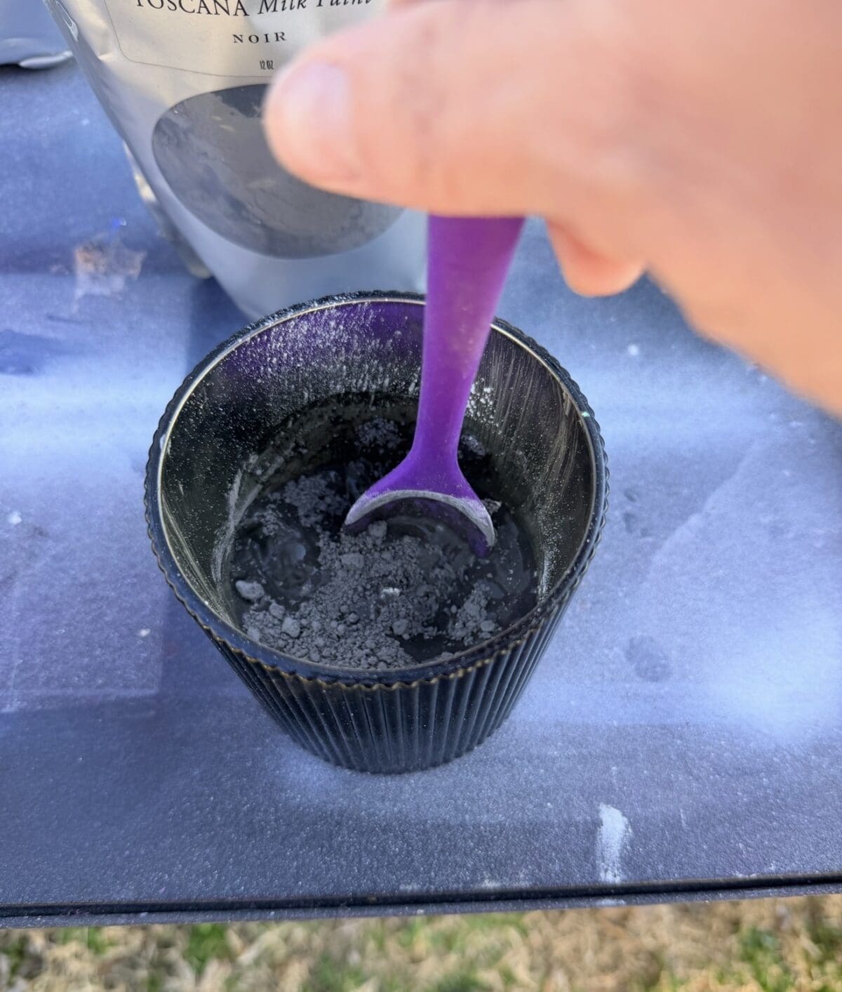 A hand stirs black powder and water in a black cup with a purple spoon. A bag labeled "Toscana Mix" is partially visible in the background on a blue surface.