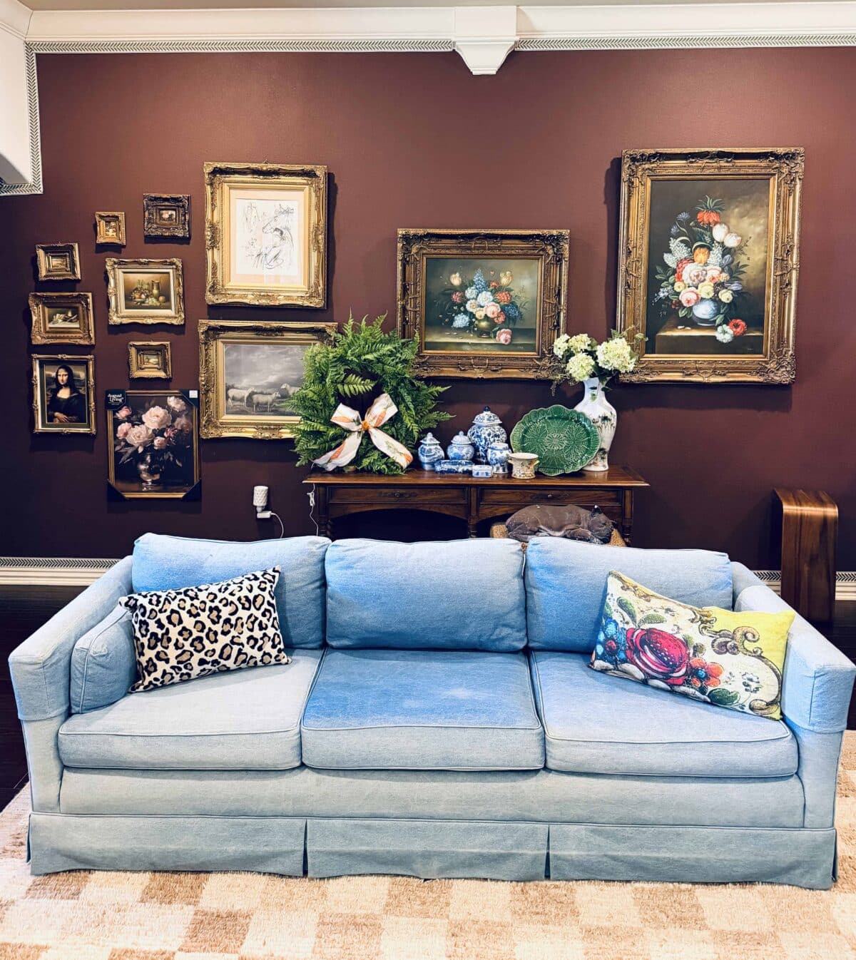 A light blue sofa with patterned pillows sits in front of a dark red wall adorned with ornate framed paintings and a wreath, perfect for a spring home tour, with floral arrangements and vases on the wooden console table adding fresh seasonal charm.