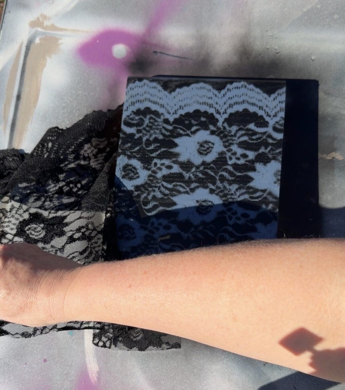 A hand holding black lace over a black canvas, partially spray-painted with white paint, creating a lace pattern. The background has splashes of pink and gray paint, and the scene is lit by sunlight, casting shadows.
