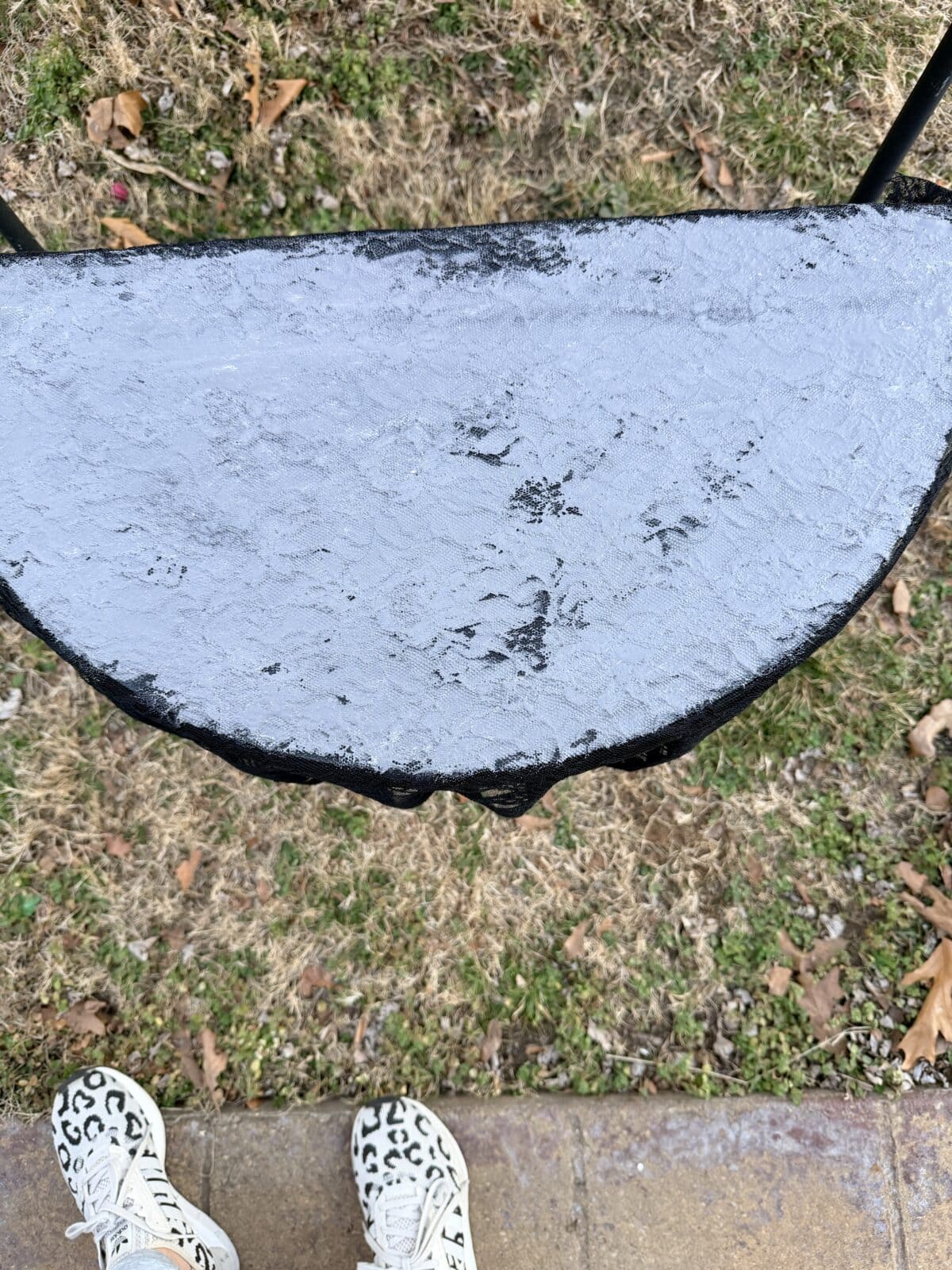 Half-circle swing seat covered in a layer of frost or ice, hanging above dry grass and scattered leaves. Two feet in white shoes with black leopard print are visible at the bottom of the image.