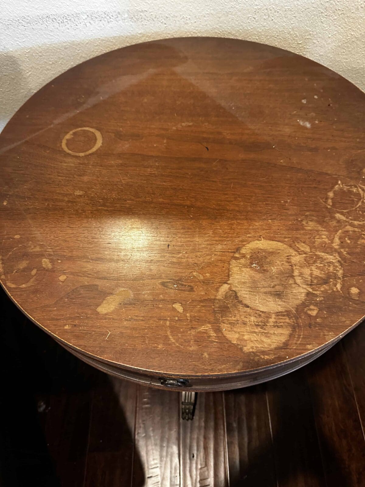 A round wooden table with several visible water stains and rings, showing marks from glasses or cups, against a light-colored wall and dark wood floor.