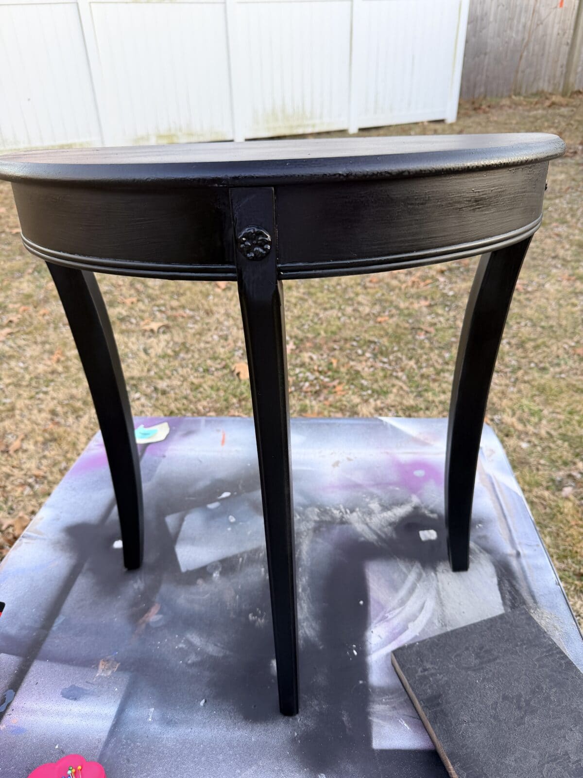 A small, half-moon-shaped black table with three legs stands on a spray-painted surface outdoors, with a white fence and dry grass in the background.