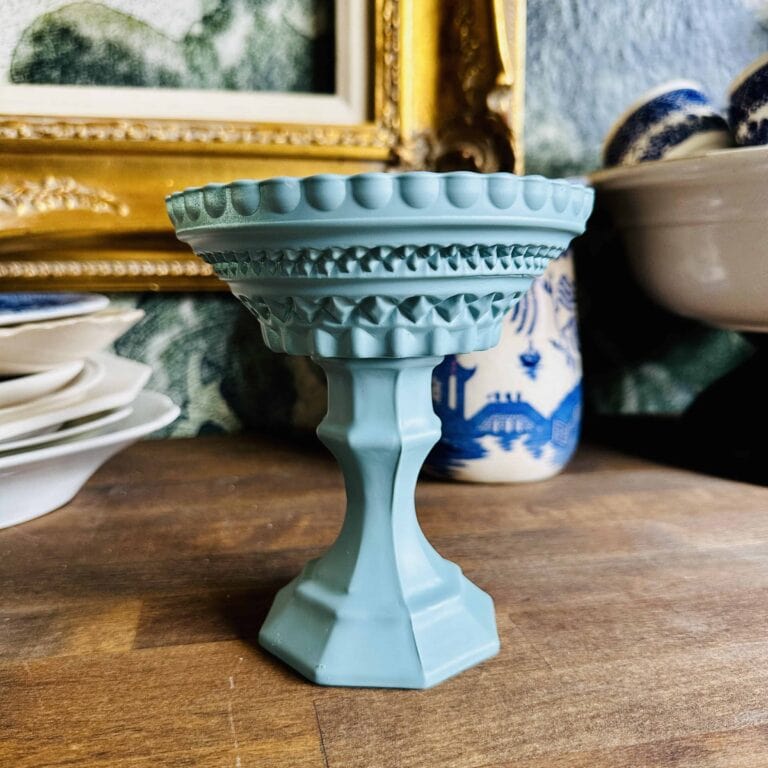 A blue, ornate ceramic pedestal bowl with intricate geometric patterns sits on a wooden surface. It's surrounded by vintage dishes and a decorative golden frame in the background.