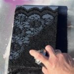A hand is holding a spray paint can, spraying over a piece of lace fabric placed on top of a rectangular object, likely to create a decorative pattern. The background has splashes of pink, white, and gray.