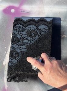 A hand is holding a spray paint can, spraying over a piece of lace fabric placed on top of a rectangular object, likely to create a decorative pattern. The background has splashes of pink, white, and gray.