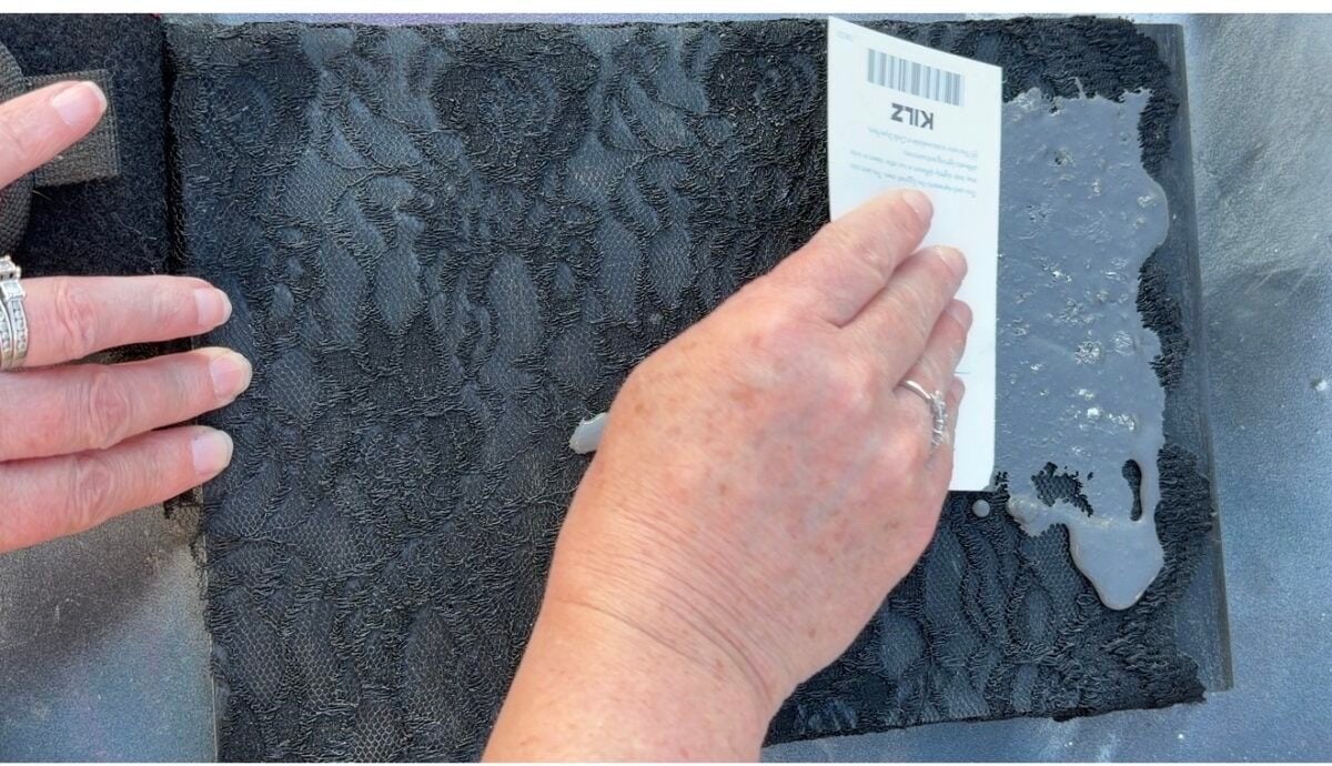 A person's hands spread a white, viscous substance over black lace fabric using a white plastic card. The lace is on a flat surface, and one hand holds the card while the other secures the fabric.