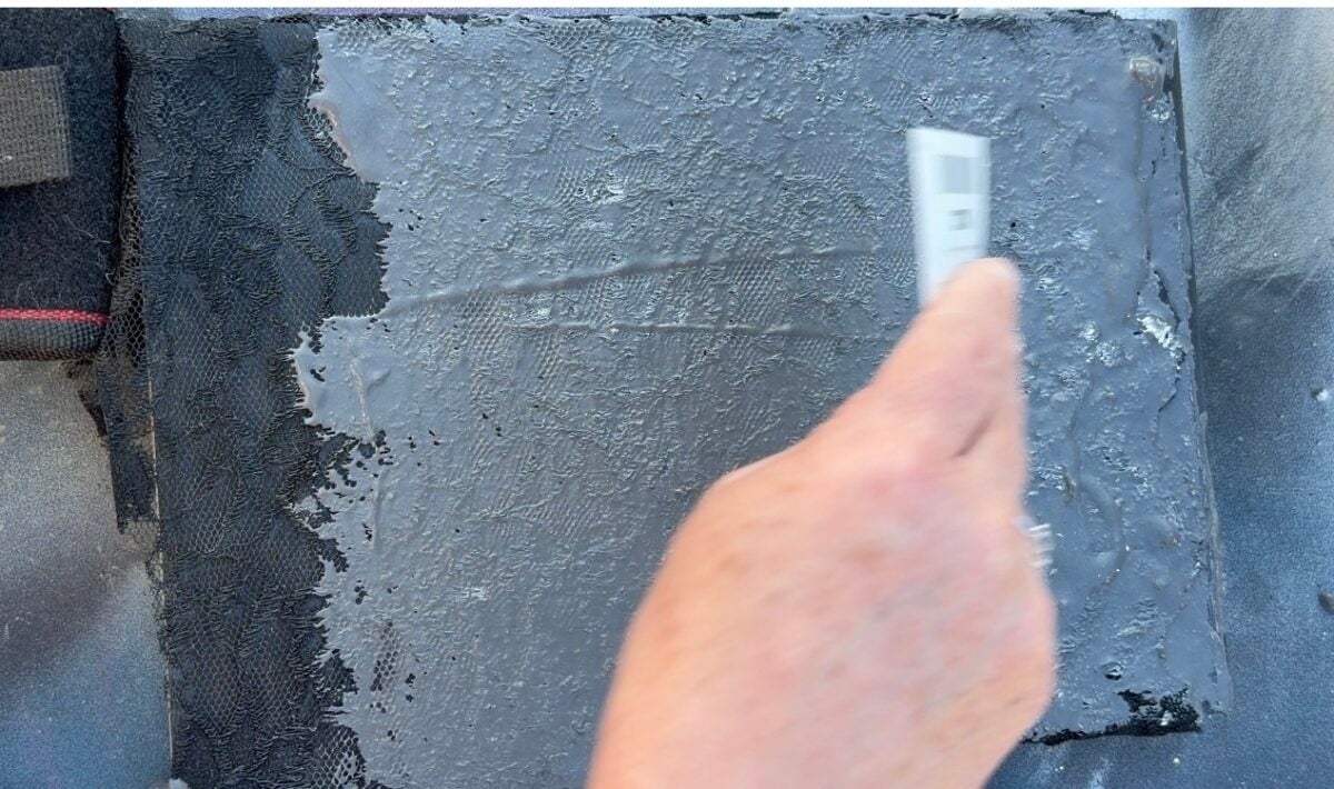 A hand uses a metal tool to smooth or scrape a black, textured surface, possibly applying or removing a coating on a rectangular object.