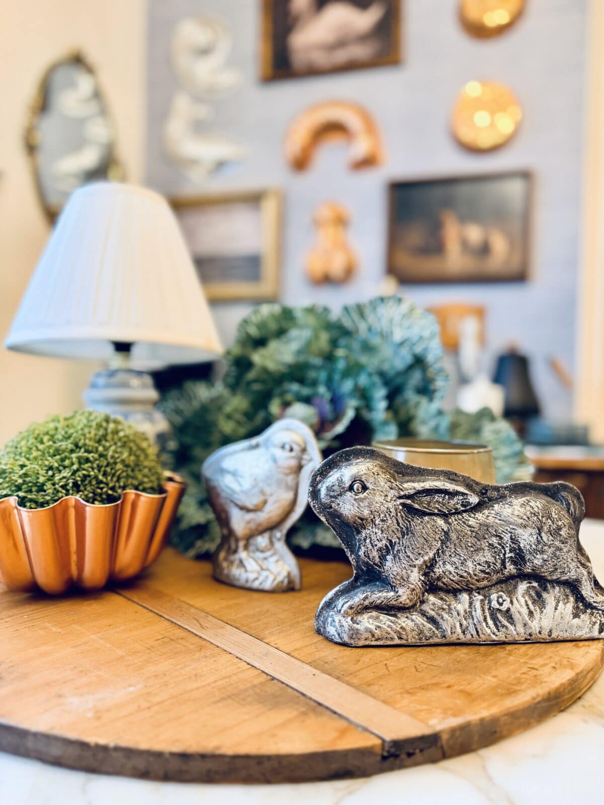 A round wooden tray displays a small green plant in a copper pot, a bird and a rabbit figurine, and some greenery—perfect for a spring home tour. In the background, there’s a white lamp and a wall decorated with pictures and art.