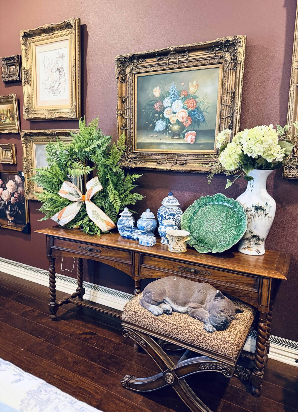 A cozy interior styled for a spring home tour features ornate wall art, a wooden console table with blue-and-white ceramics, a green leaf plate, hydrangeas, and a fern. A dog naps on a cushioned bench beneath the table.