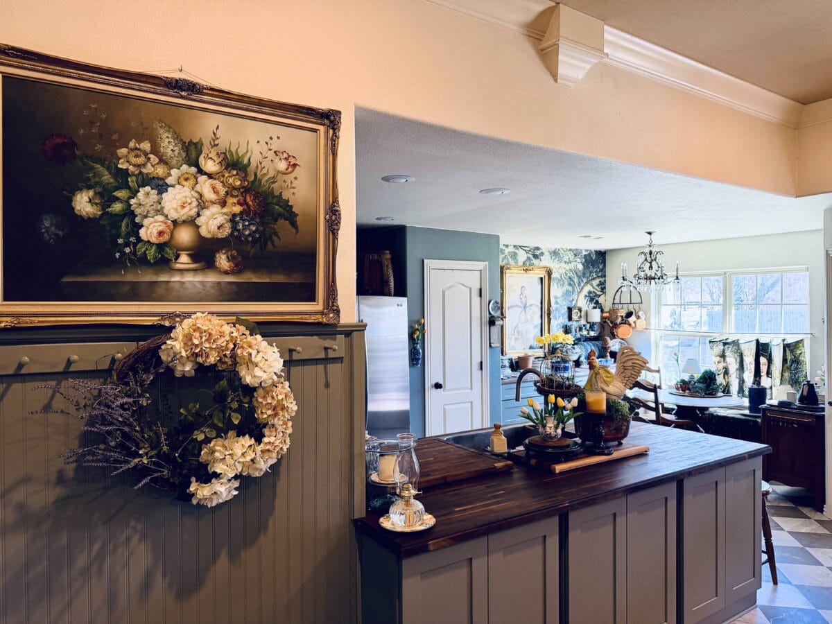 A cozy kitchen with a wooden island, decorated with flowers and a rooster statue, sets the scene for a spring home tour. A floral wreath and painting adorn the wall, while the dining area features large windows and a chandelier in the background.