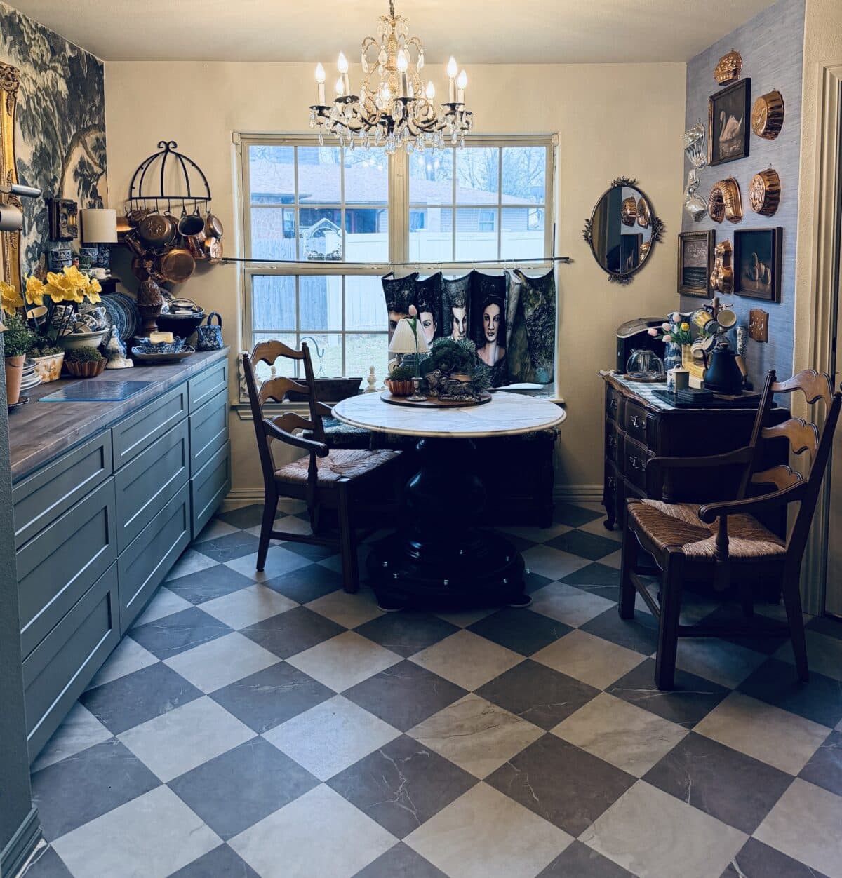 A cozy dining room with checkered tile floor, a round table with chairs, blue cabinets, vintage decor, and a chandelier welcomes you. Perfect for a spring home tour, it features natural light, photos, and plants on the surfaces and walls.