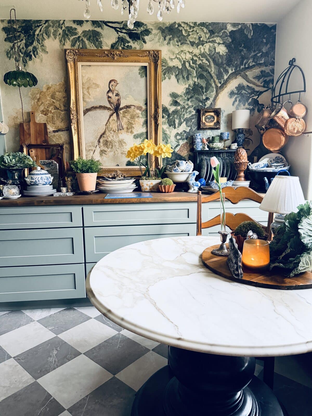 A cozy kitchen with green cabinets, a marble-topped round table, potted plants, decorative dishes, copper pots, and a large framed bird painting on a leafy mural wall—perfect for your spring home tour. Natural light fills the space.