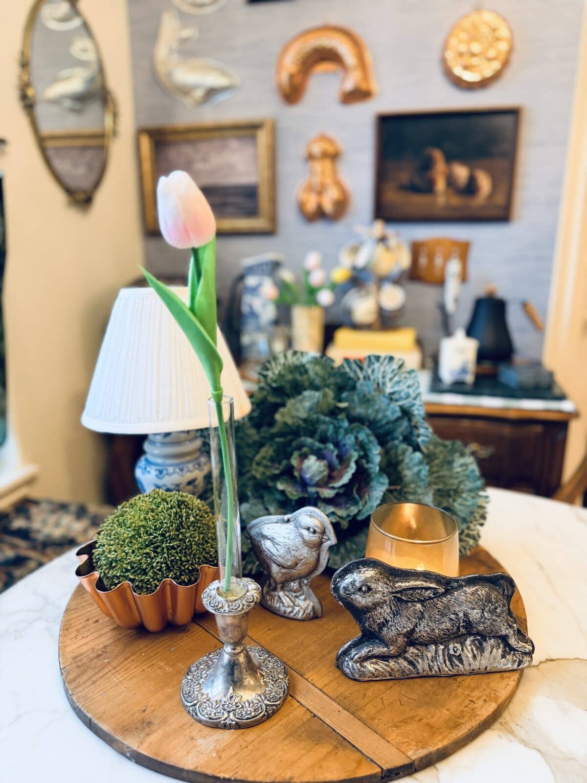 A marble table displays a pink tulip in a vase, potted moss, a decorative cabbage, a candle, and two rabbit figurines—perfect touches for your spring home tour. In the background, framed art and gold wall decor hang on a blue wall.