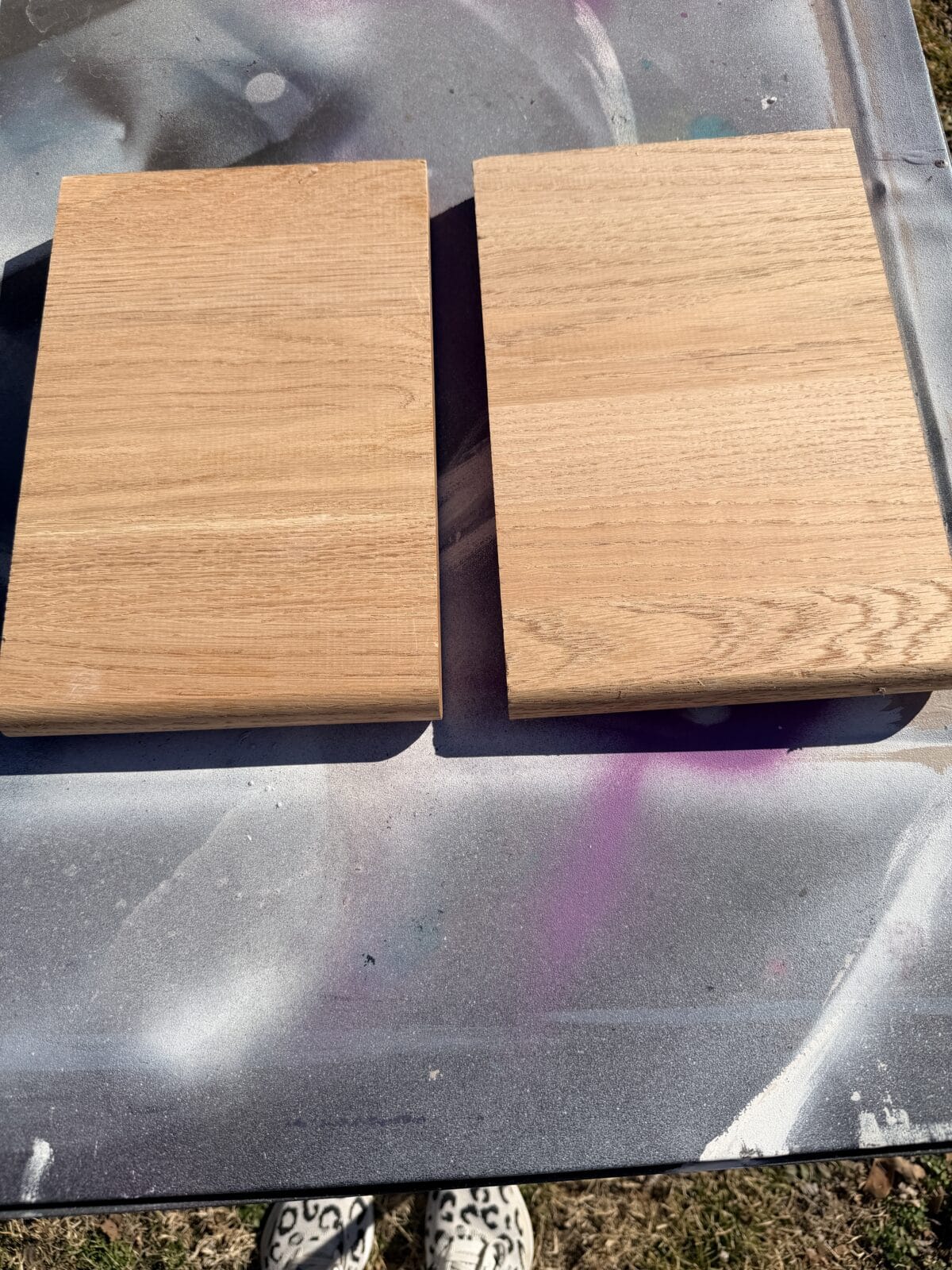 Two rectangular wooden boards sit side by side on a surface with spray paint splatters in gray, purple, and white. The boards appear unfinished, and the tips of shoes are visible at the bottom of the image.