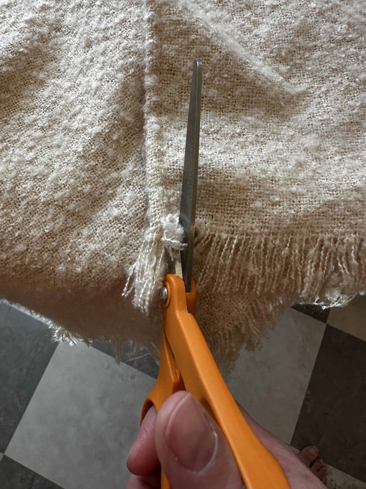 A hand uses orange-handled scissors to cut the fringe of a textured beige fabric on a surface with a checkered floor in the background.