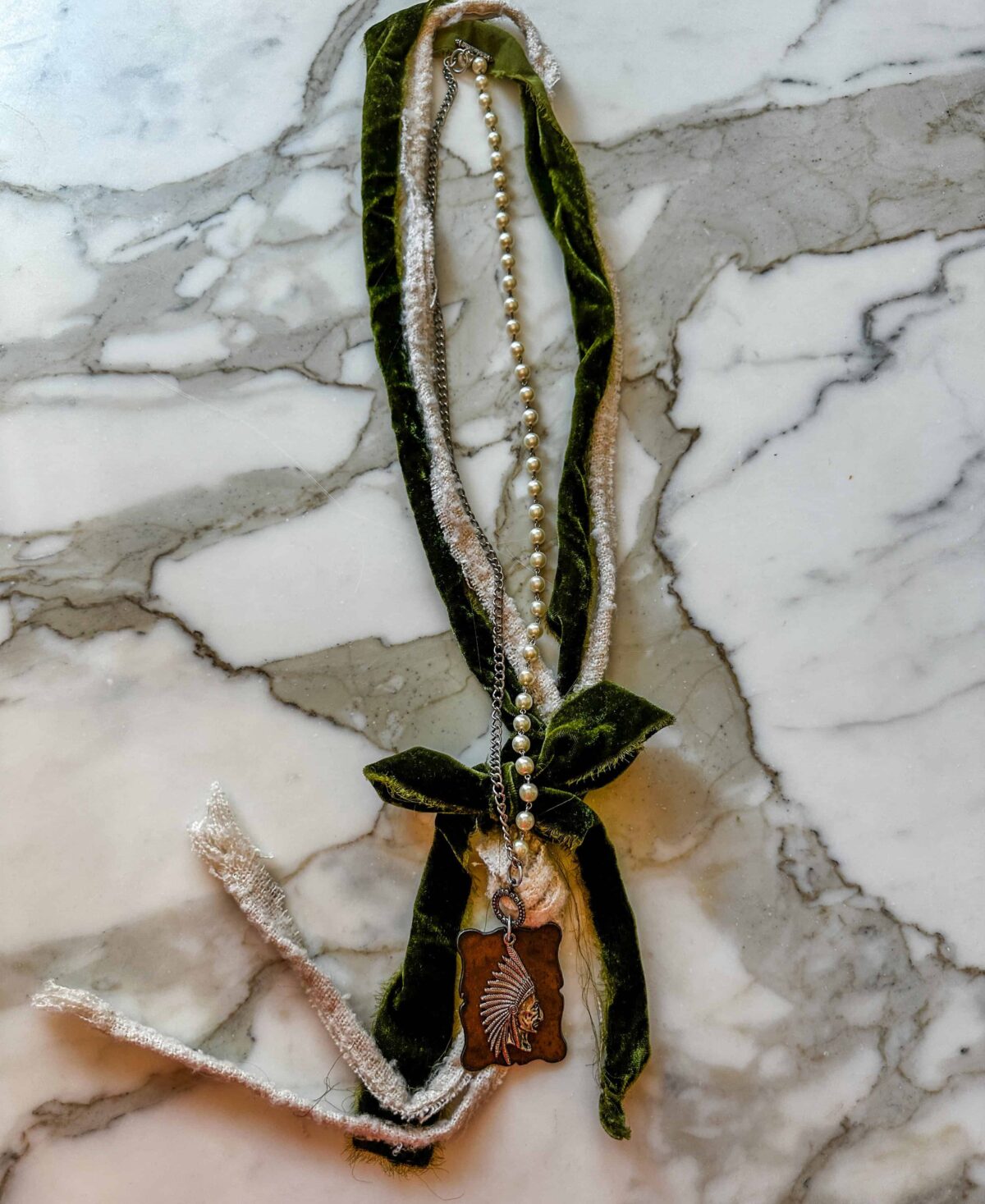 A long, green velvet ribbon and a string of pearls are tied together, adorned with a pendant featuring a delicate leaf design, all resting on a white and gray marble surface.