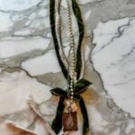 A long, green velvet ribbon and a string of pearls are tied together, adorned with a pendant featuring a delicate leaf design, all resting on a white and gray marble surface.
