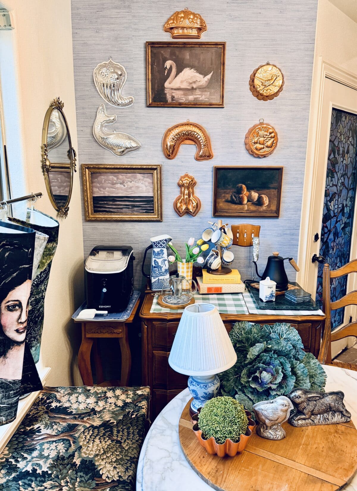A cozy room with a desk and table, decorated for a spring home tour—vintage art, copper molds, and plants abound. Framed paintings and fish-shaped molds hang above a cluttered desk filled with books, a lamp, and various items.