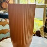 A tall, ribbed copper vase sits on a marble table in a sunlit kitchen, with a window and hanging pots visible in the background.