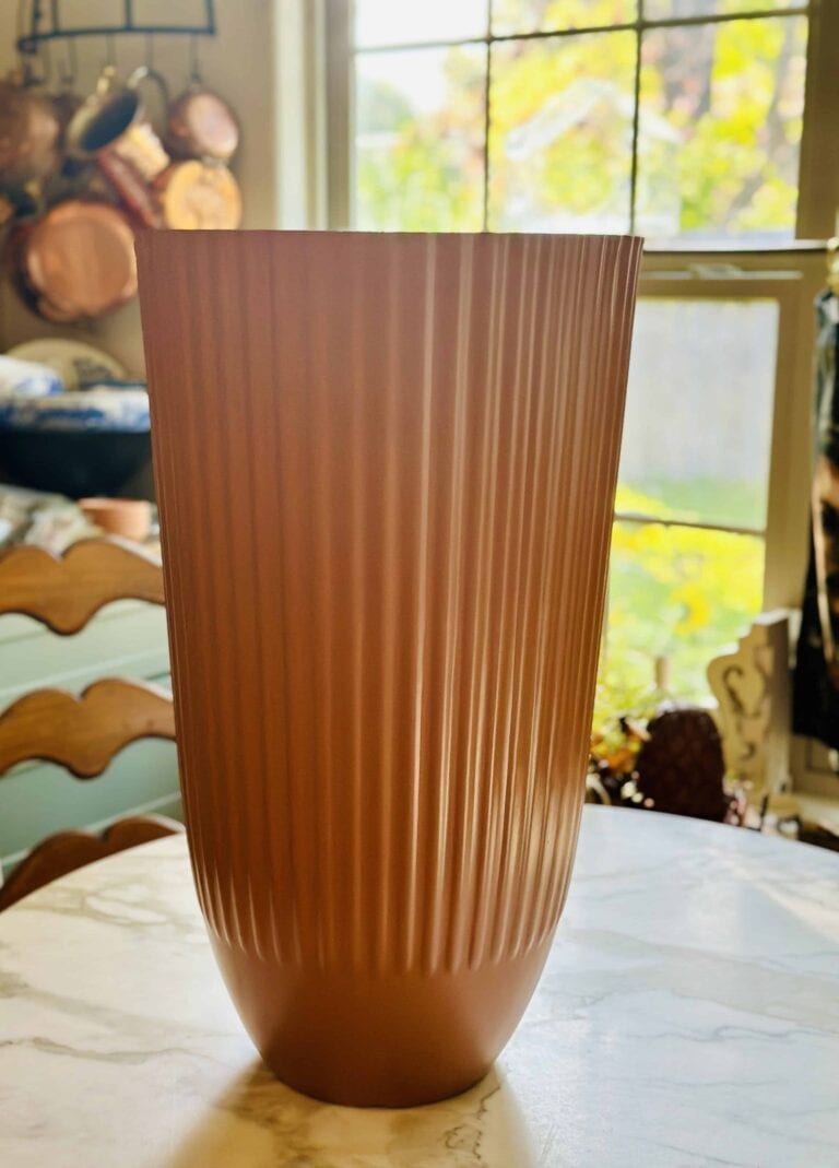 A tall, ribbed copper vase sits on a marble table in a sunlit kitchen, with a window and hanging pots visible in the background.
