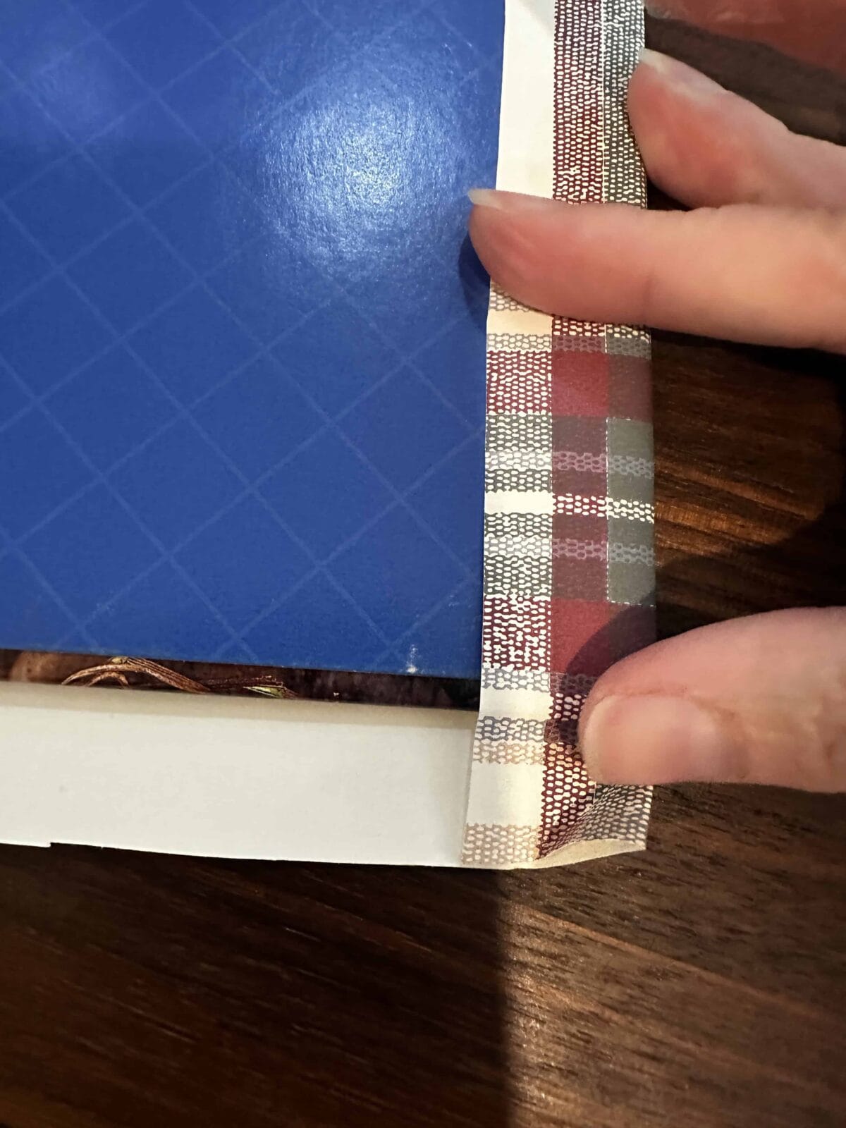 A hand peels back a strip of patterned tape from the edge of a blue, grid-patterned book cover resting on a wooden surface.
